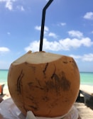 A fresh coconut with a straw inserted in the top, set against a bright, clear sky and turquoise ocean in the background.