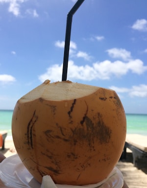 A fresh coconut with a straw inserted in the top, set against a bright, clear sky and turquoise ocean in the background.