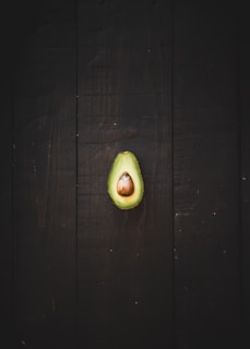 A halved avocado with a large central pit sits on a dark wooden surface. The vibrant green of the avocado contrasts with the deep brown background, emphasizing the smooth texture of the fruit.