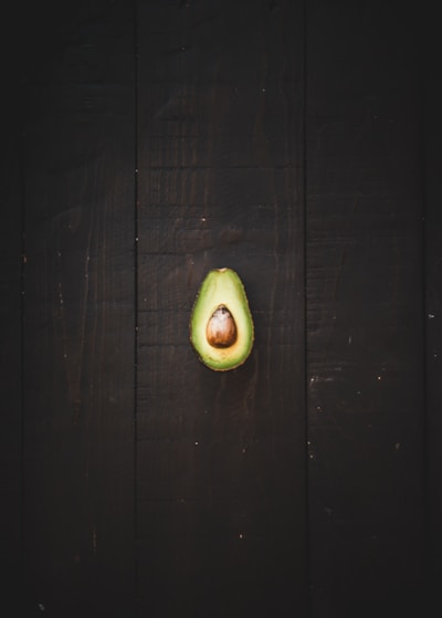 A halved avocado with a large central pit sits on a dark wooden surface. The vibrant green of the avocado contrasts with the deep brown background, emphasizing the smooth texture of the fruit.
