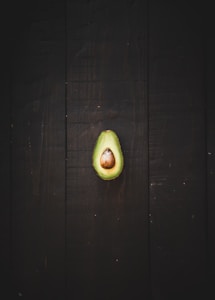 A halved avocado with a large central pit sits on a dark wooden surface. The vibrant green of the avocado contrasts with the deep brown background, emphasizing the smooth texture of the fruit.