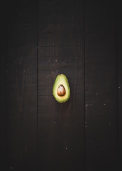 A halved avocado with a large central pit sits on a dark wooden surface. The vibrant green of the avocado contrasts with the deep brown background, emphasizing the smooth texture of the fruit.