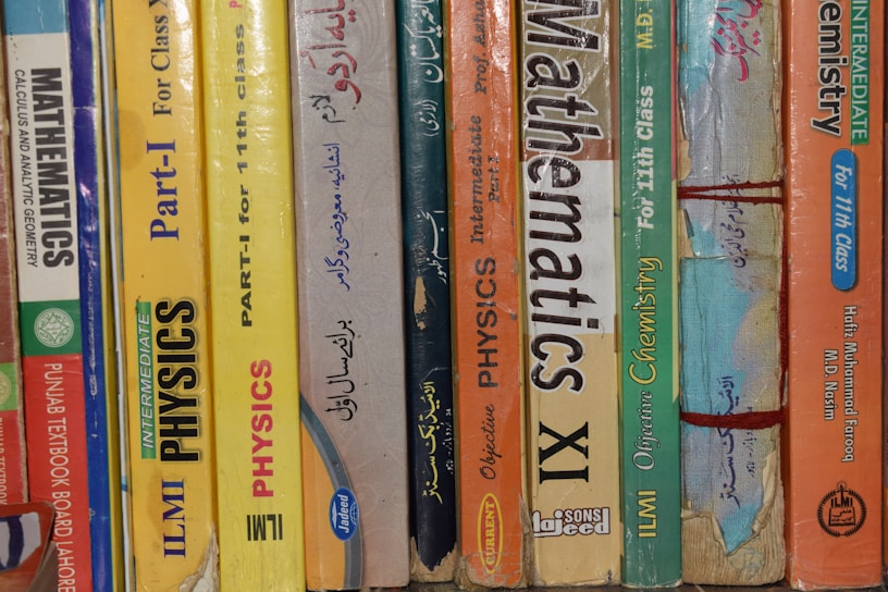 A row of textbooks stacked vertically on a shelf, with subjects like mathematics, physics, and chemistry. The books vary in color and are written in both English and another language. The spines display titles, grades, and authors, indicating they are for 11th grade classes.