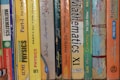 A row of textbooks stacked vertically on a shelf, with subjects like mathematics, physics, and chemistry. The books vary in color and are written in both English and another language. The spines display titles, grades, and authors, indicating they are for 11th grade classes.