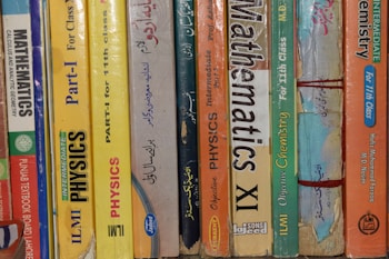 A row of textbooks stacked vertically on a shelf, with subjects like mathematics, physics, and chemistry. The books vary in color and are written in both English and another language. The spines display titles, grades, and authors, indicating they are for 11th grade classes.