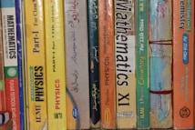 A row of textbooks stacked vertically on a shelf, with subjects like mathematics, physics, and chemistry. The books vary in color and are written in both English and another language. The spines display titles, grades, and authors, indicating they are for 11th grade classes.