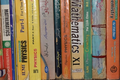 A row of textbooks stacked vertically on a shelf, with subjects like mathematics, physics, and chemistry. The books vary in color and are written in both English and another language. The spines display titles, grades, and authors, indicating they are for 11th grade classes.