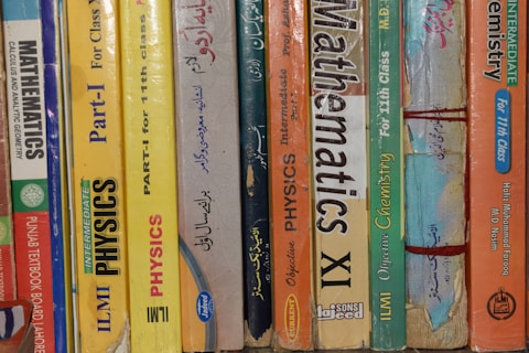 A row of textbooks stacked vertically on a shelf, with subjects like mathematics, physics, and chemistry. The books vary in color and are written in both English and another language. The spines display titles, grades, and authors, indicating they are for 11th grade classes.