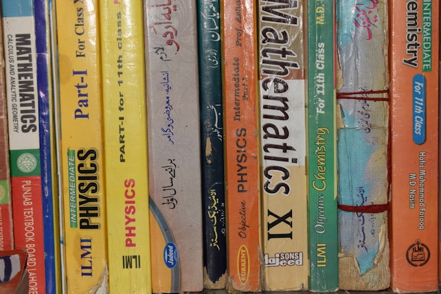 A row of textbooks stacked vertically on a shelf, with subjects like mathematics, physics, and chemistry. The books vary in color and are written in both English and another language. The spines display titles, grades, and authors, indicating they are for 11th grade classes.