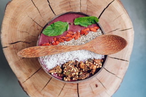 A bowl filled with a smoothie is topped with vibrant ingredients such as mint leaves, chia seeds, goji berries, coconut shreds, and granola. It is presented on a wooden surface with a textured wooden spoon resting on top.