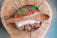 A bowl filled with a smoothie is topped with vibrant ingredients such as mint leaves, chia seeds, goji berries, coconut shreds, and granola. It is presented on a wooden surface with a textured wooden spoon resting on top.