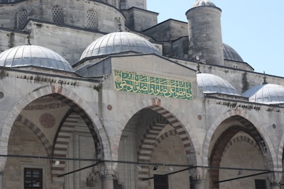 A historic stone building with domed roofs and arched facades featuring intricate carvings and patterns. There is an ornate green and gold Arabic inscription on the front section. The architecture reflects traditional Islamic design, with detailed motifs and symmetrical arches.