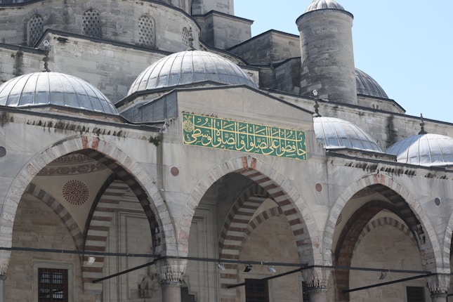 A historic stone building with domed roofs and arched facades featuring intricate carvings and patterns. There is an ornate green and gold Arabic inscription on the front section. The architecture reflects traditional Islamic design, with detailed motifs and symmetrical arches.