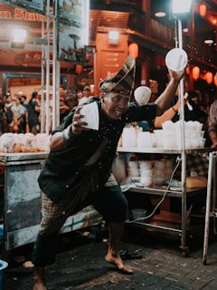 A street performer energetically entertains a crowd by skillfully tossing food containers. He's wearing traditional attire, and the scene takes place at a vibrant night market, warmly lit by overhead lights and colorful lanterns. The audience appears engaged and amused, adding to the lively atmosphere.