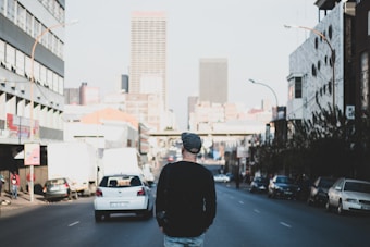 A person wearing a beanie and a black sweater walks down a city street. The street is lined with cars and tall buildings on either side, with skyscrapers visible in the distance. The atmosphere is urban and somewhat bustling.