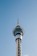 A tall, modern telecommunications tower with a sleek design stands against a clear blue sky. The structure features a circular observation deck near the top, where the tower's glass windows reflect light. The tower tapers upwards with a needle-like antenna protruding from the top.