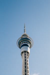 A tall, modern telecommunications tower with a sleek design stands against a clear blue sky. The structure features a circular observation deck near the top, where the tower's glass windows reflect light. The tower tapers upwards with a needle-like antenna protruding from the top.