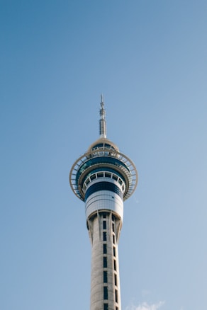A tall, modern telecommunications tower with a sleek design stands against a clear blue sky. The structure features a circular observation deck near the top, where the tower's glass windows reflect light. The tower tapers upwards with a needle-like antenna protruding from the top.