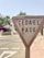 A triangular traffic sign with the text 'CEDA EL PASO' is prominently displayed. The sign appears to be aged, with noticeable dirt marks. It is situated on the side of a paved road, with a sidewalk next to it. In the background, there are flowering bushes and trees, partially blurred, suggesting a residential or suburban area.