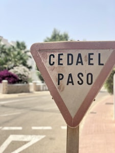 A triangular traffic sign with the text 'CEDA EL PASO' is prominently displayed. The sign appears to be aged, with noticeable dirt marks. It is situated on the side of a paved road, with a sidewalk next to it. In the background, there are flowering bushes and trees, partially blurred, suggesting a residential or suburban area.