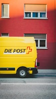 A bright yellow delivery van with the words 'DIE POST' in black and a red cross logo is parked on a street in front of a red building. The building has several windows with closed shutters.