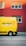A bright yellow delivery van with the words 'DIE POST' in black and a red cross logo is parked on a street in front of a red building. The building has several windows with closed shutters.