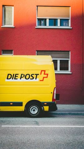 A bright yellow delivery van with the words 'DIE POST' in black and a red cross logo is parked on a street in front of a red building. The building has several windows with closed shutters.