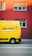 A bright yellow delivery van with the words 'DIE POST' in black and a red cross logo is parked on a street in front of a red building. The building has several windows with closed shutters.