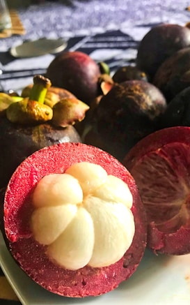 A close-up of several mangosteens, with one prominently cut in half to show its white segmented fruit flesh. The fruits have a dark purple rind and small green stems. The lighting creates a warm, inviting atmosphere.