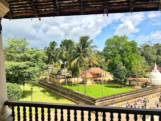 A lush, green courtyard with several tall palm trees, surrounded by a stone wall. To the right, a white stupa with a round base and pointed top stands prominently. In the background, a cluster of traditional buildings with red-tiled roofs is visible. The sky is partly cloudy, creating areas of both sunlight and shade across the scene.