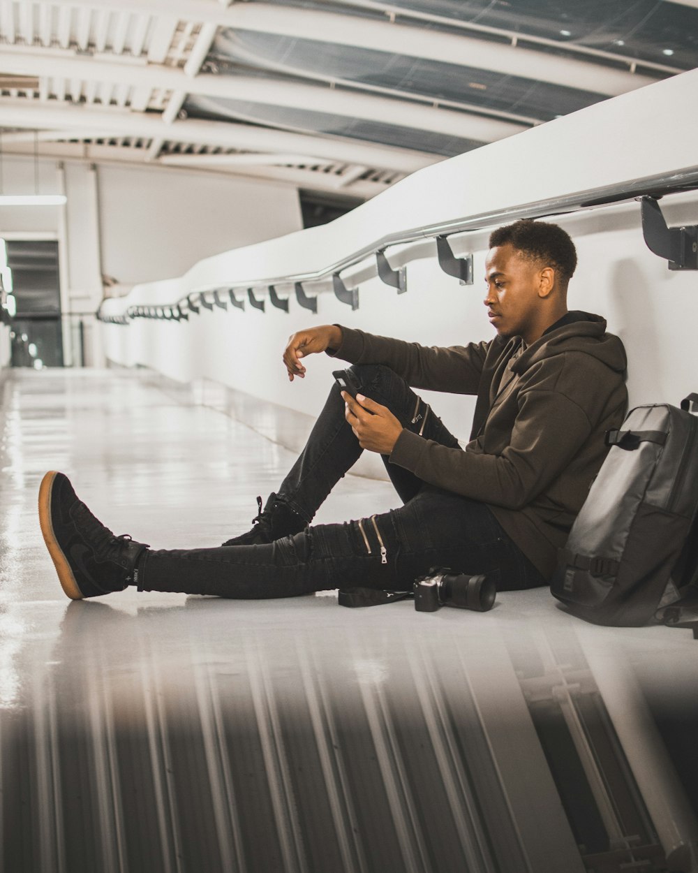 man wearing black jacket sitting while leaning on wall and using smartphone