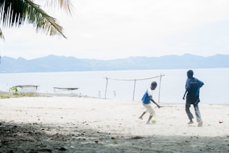 Two children play soccer on a sandy beach, with one kicking a ball towards the other. Palm leaves hang overhead, and in the background, a serene body of water and distant mountains create a peaceful landscape. A simple makeshift goalpost is present, and a couple of small boats are visible on the water.