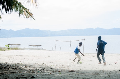 Two children play soccer on a sandy beach, with one kicking a ball towards the other. Palm leaves hang overhead, and in the background, a serene body of water and distant mountains create a peaceful landscape. A simple makeshift goalpost is present, and a couple of small boats are visible on the water.