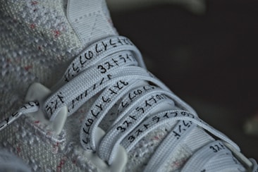 Close-up of a white sneaker with textured fabric and shoelaces inscribed with black characters. The characters appear to be Japanese or Korean, and the laces are neatly tied in a standard crisscross pattern.