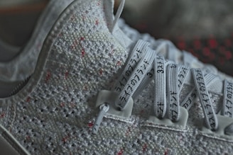 A close-up view of a sneaker featuring a textured knit upper with red and white patterns. The laces are prominently visible, marked with black text in a non-English script, possibly Japanese or Chinese.