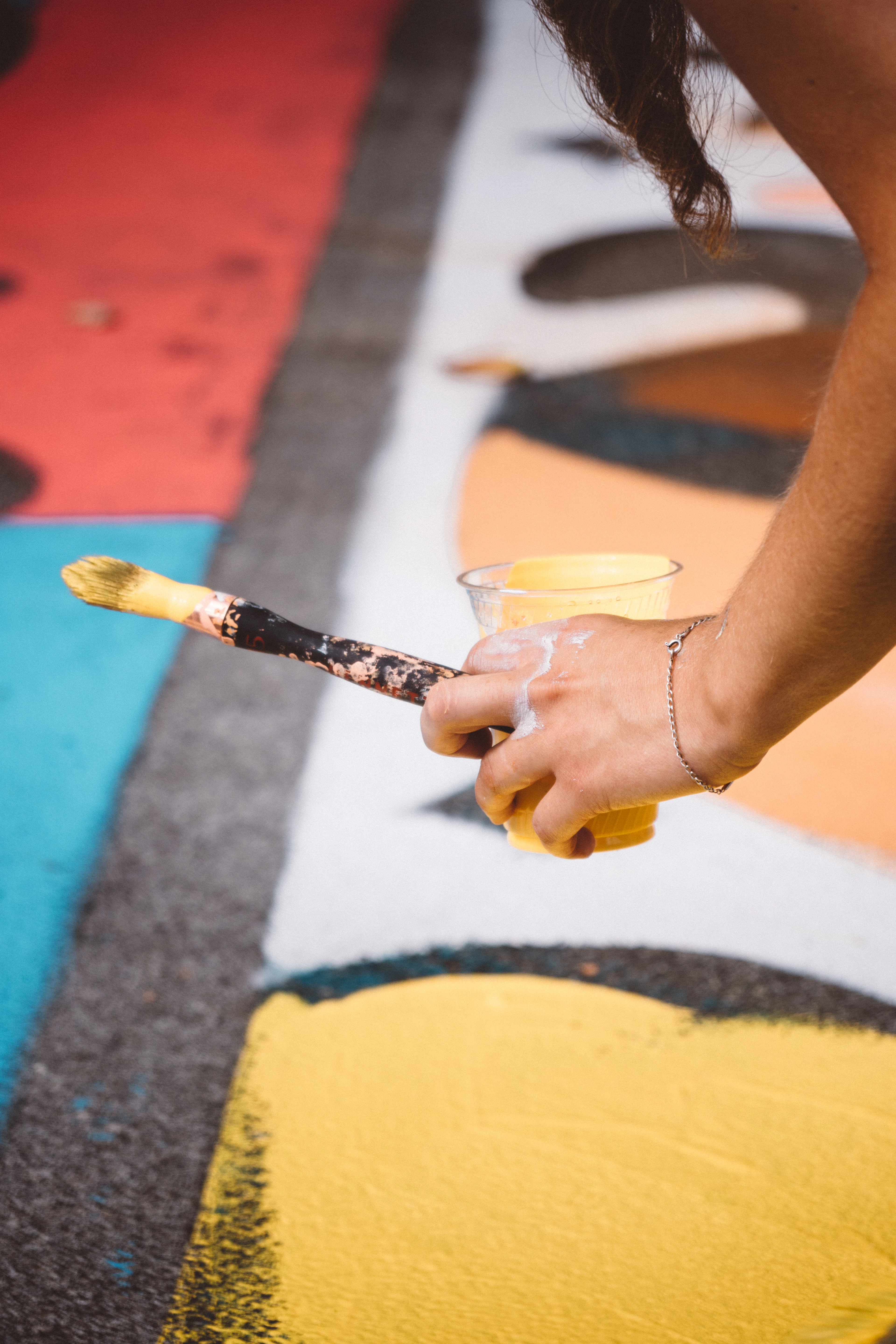 A hand holding a cup of yellow paint and a brush in front of a mural.