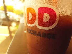 A plastic cup with droplets on its surface, featuring a logo with the letters 'DD' in bold colors. The background appears to be a blurred outdoor setting with sunlight filtering through, highlighting the cup and creating a warm ambiance.