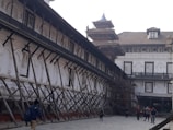 An old architectural building with traditional design, supported by wooden structures angled for reinforcement. The building features arched windows with shutters, and there are people walking in the paved courtyard. The structure appears to be undergoing restoration or protection.