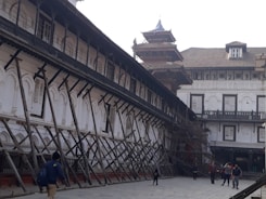 An old architectural building with traditional design, supported by wooden structures angled for reinforcement. The building features arched windows with shutters, and there are people walking in the paved courtyard. The structure appears to be undergoing restoration or protection.