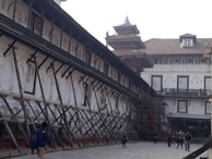 An old architectural building with traditional design, supported by wooden structures angled for reinforcement. The building features arched windows with shutters, and there are people walking in the paved courtyard. The structure appears to be undergoing restoration or protection.