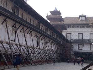 An old architectural building with traditional design, supported by wooden structures angled for reinforcement. The building features arched windows with shutters, and there are people walking in the paved courtyard. The structure appears to be undergoing restoration or protection.