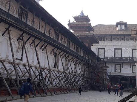 An old architectural building with traditional design, supported by wooden structures angled for reinforcement. The building features arched windows with shutters, and there are people walking in the paved courtyard. The structure appears to be undergoing restoration or protection.