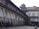 An old architectural building with traditional design, supported by wooden structures angled for reinforcement. The building features arched windows with shutters, and there are people walking in the paved courtyard. The structure appears to be undergoing restoration or protection.