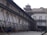 An old architectural building with traditional design, supported by wooden structures angled for reinforcement. The building features arched windows with shutters, and there are people walking in the paved courtyard. The structure appears to be undergoing restoration or protection.