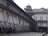An old architectural building with traditional design, supported by wooden structures angled for reinforcement. The building features arched windows with shutters, and there are people walking in the paved courtyard. The structure appears to be undergoing restoration or protection.