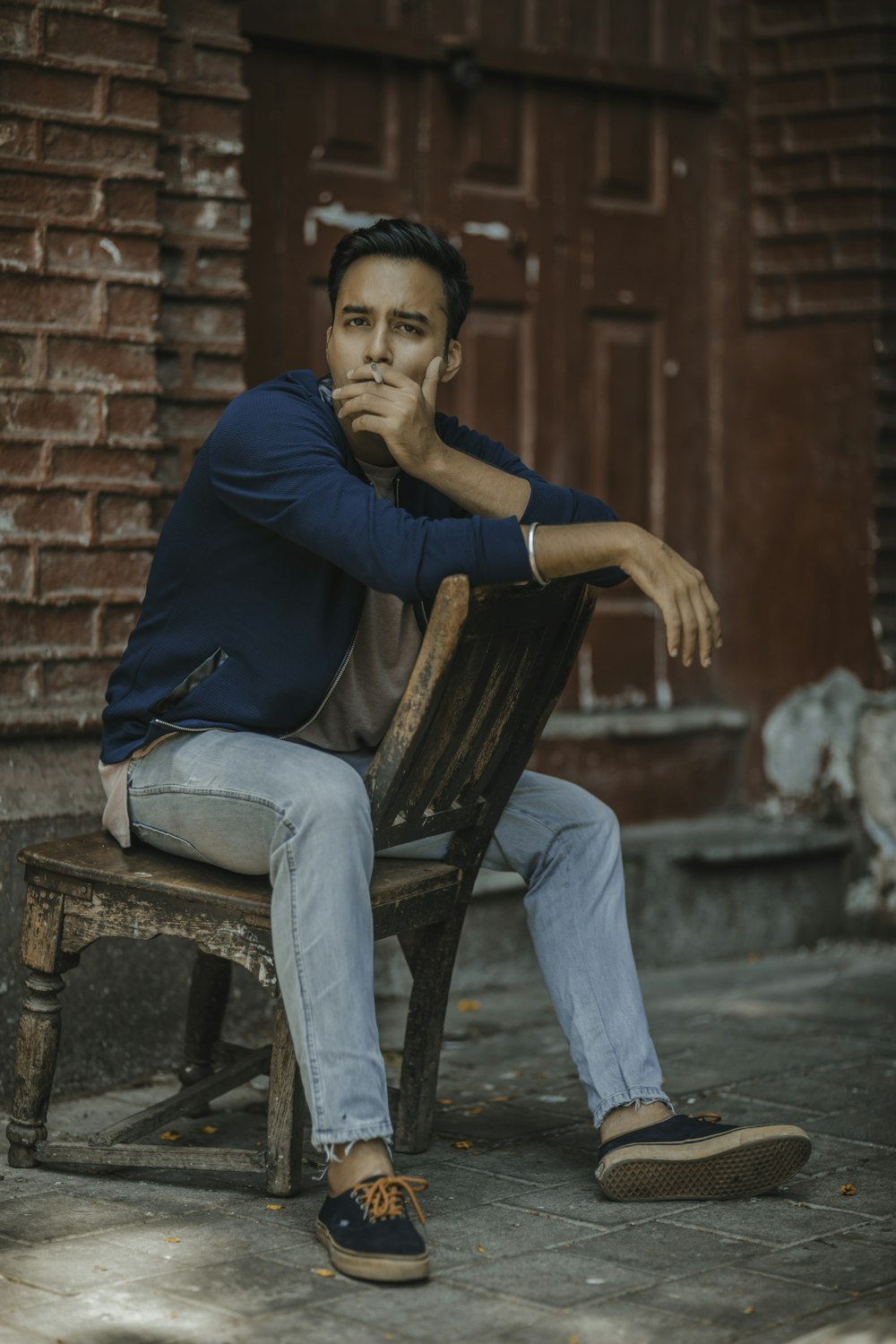 man in blue jacket smoking while sitting on chair near building