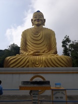 A large golden statue of a seated figure with distinct robes, positioned in front of a light sky. Below the statue, there is a donation box with signs written in multiple languages. Surrounding the statue are trees, adding a natural backdrop.