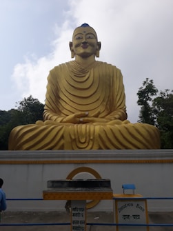 A large golden statue of a seated figure with distinct robes, positioned in front of a light sky. Below the statue, there is a donation box with signs written in multiple languages. Surrounding the statue are trees, adding a natural backdrop.