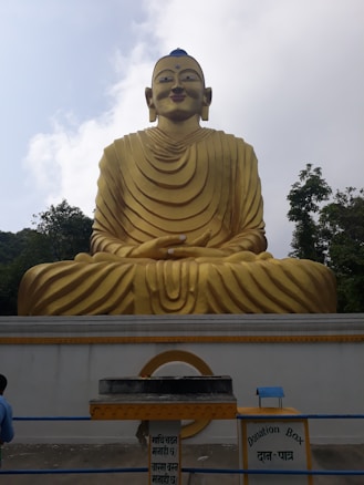A large golden statue of a seated figure with distinct robes, positioned in front of a light sky. Below the statue, there is a donation box with signs written in multiple languages. Surrounding the statue are trees, adding a natural backdrop.
