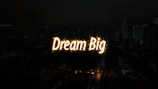 A nighttime cityscape with tall buildings in the background. The words 'Dream Big' are illuminated in bright, glowing letters across the center of the image, resembling a light display or sparkler effect.